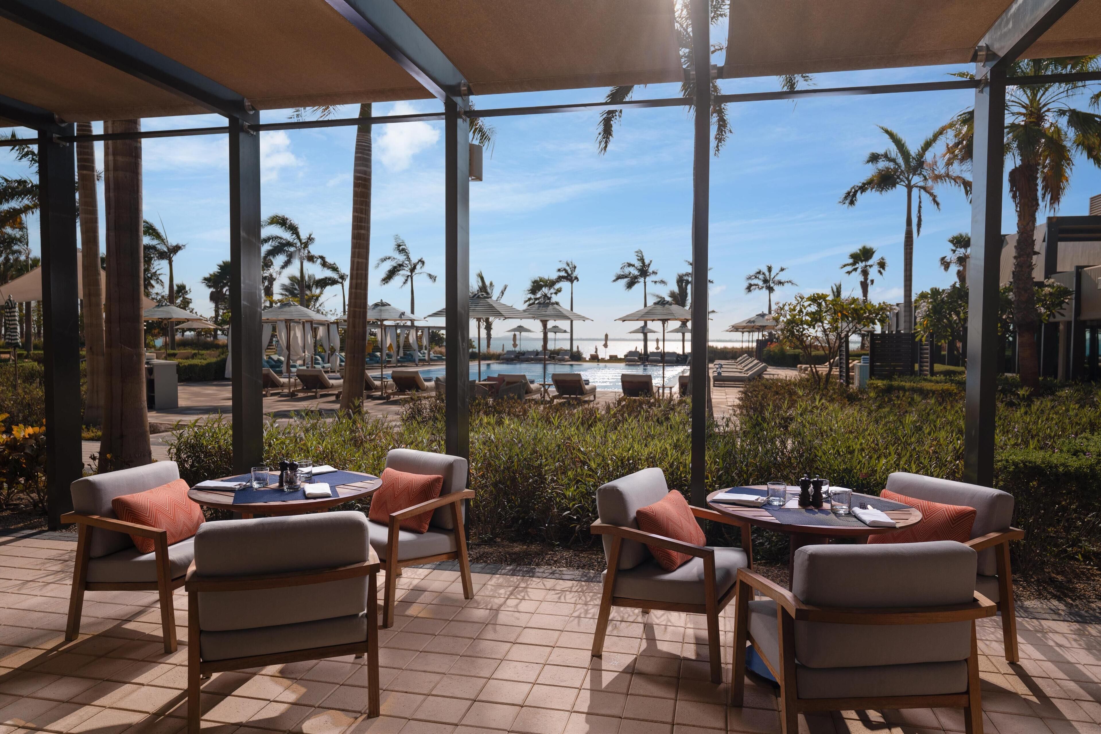 A covered outdoor seating area with tables and chairs, overlooking a pool and palm trees.