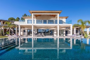 A modern two-story house with a large pool, outdoor seating, and a clear blue sky.