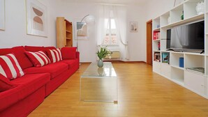 Uma sala de estar com um sofá vermelho, uma mesa de centro de vidro, uma TV sobre um suporte branco e uma estante com diversos objetos.
