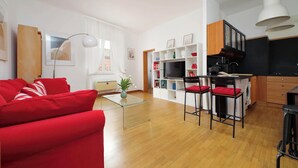 Uma sala de estar moderna com um sofá vermelho, uma mesa de centro de vidro e uma área de cozinha com televisão, banquetas de bar e armários de madeira.