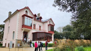 Ein pinkfarbenes Gebäude mit einem großen Wasserrad, umgeben von viel Grün und einigen Personen.