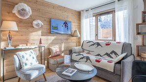 A cozy living room with a wooden wall, a sofa with a mountain pattern, a round coffee table, and a television displaying a skier. - Modern 3-bedroom apartment, ski-in/ski-out, perfect for 6 people (Huez)
