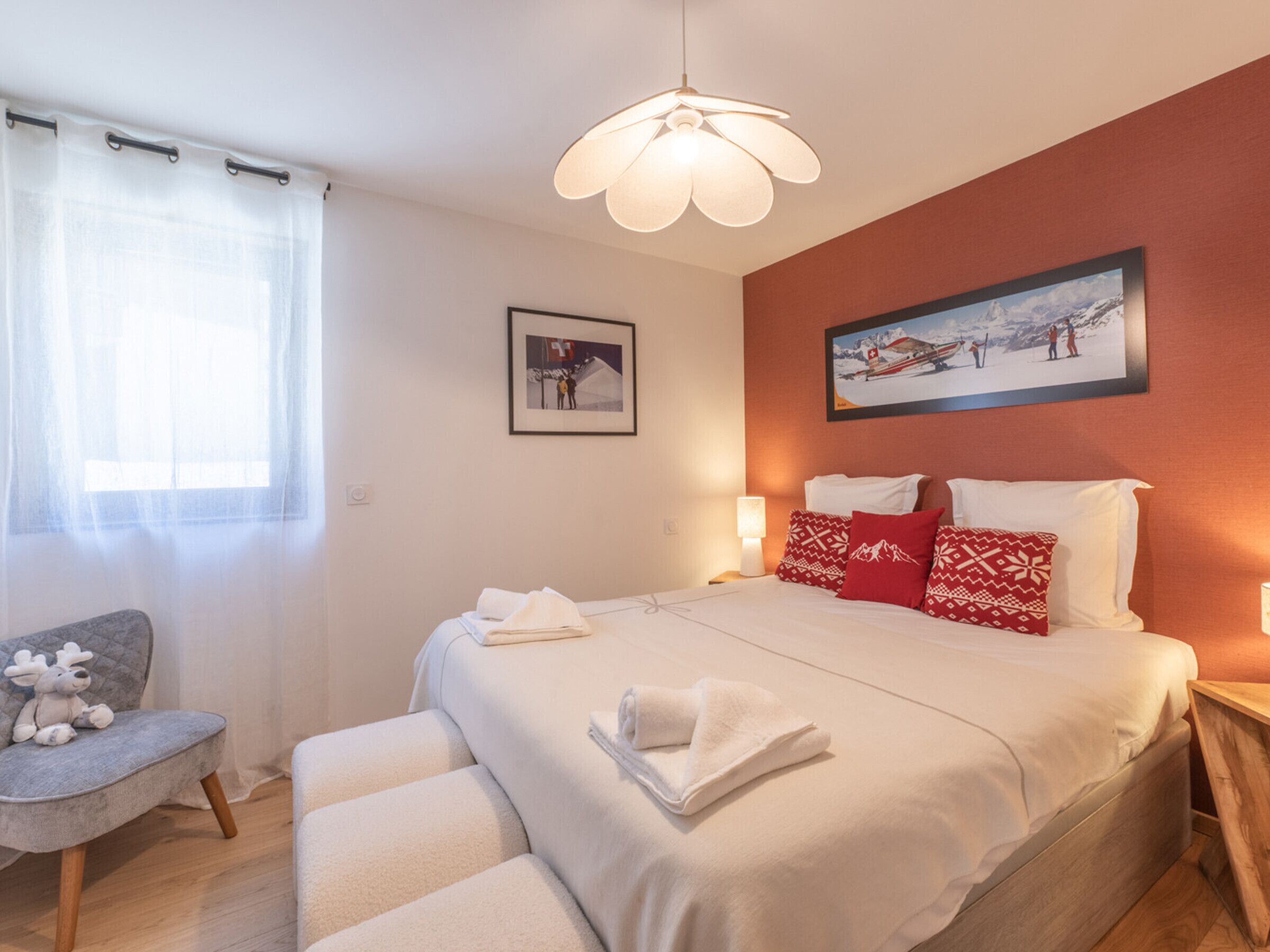 A bedroom with a bed, pillows, a bedside table, a lamp, a chair, a window with curtains, a wall-mounted picture, and a ceiling light.