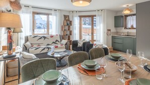 A cozy living room with a sofa, chairs, a dining table set for four, and a kitchen area. - Modern 3-bedroom apartment, ski-in/ski-out, perfect for 6 people (Huez)