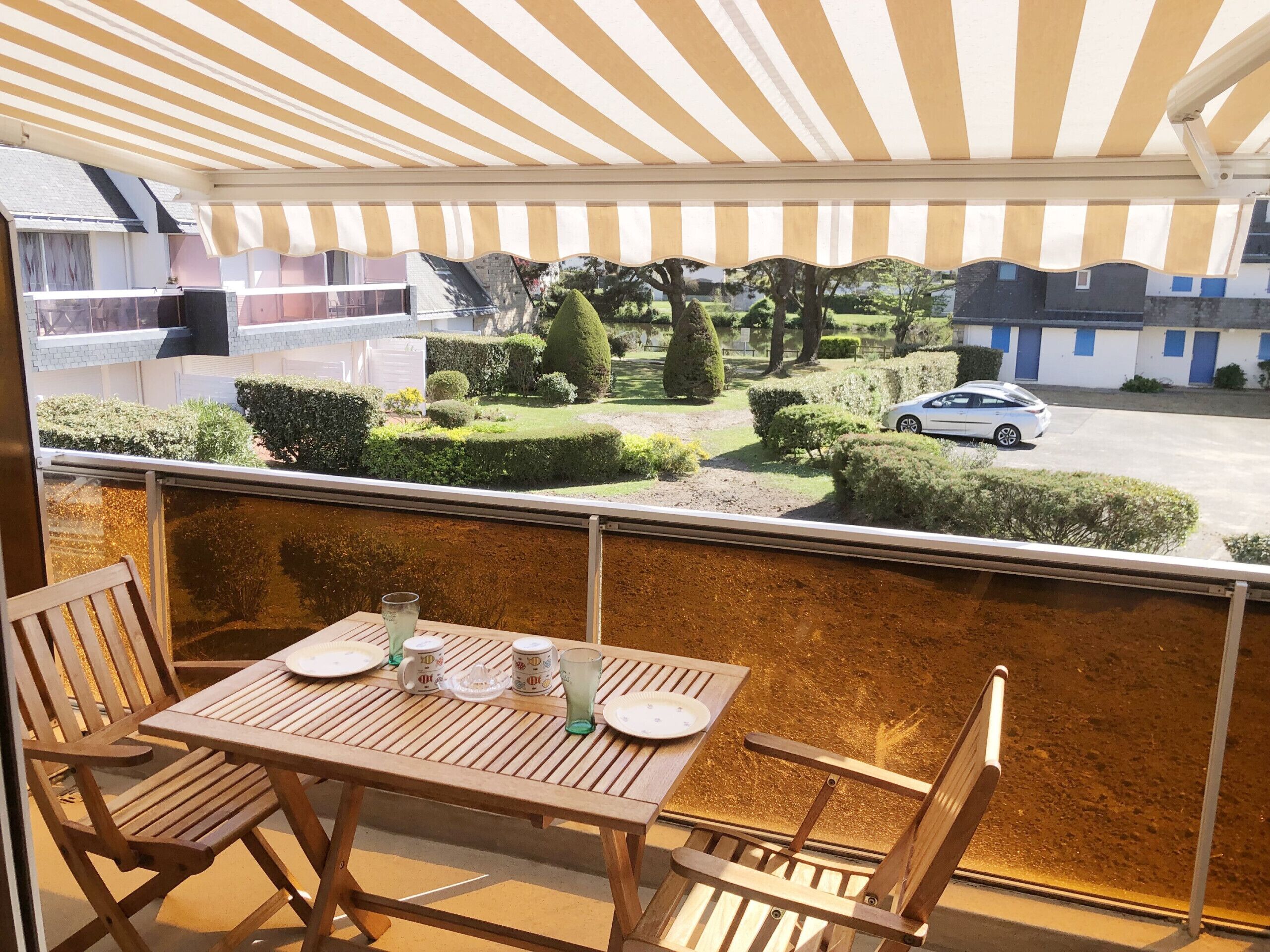 A balcony with a table set for two, overlooking a well-maintained garden and a parking area.