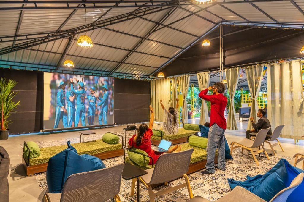 A spacious indoor area with a large screen displaying a sports event, comfortable seating areas, and a person standing with arms raised.