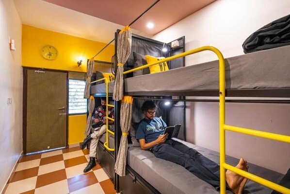 A dormitory room with bunk beds, a checkered floor, a clock on the wall, and a person reading a book. - The Hosteller Wayanad, Vythiri (Kunnathidavaka)