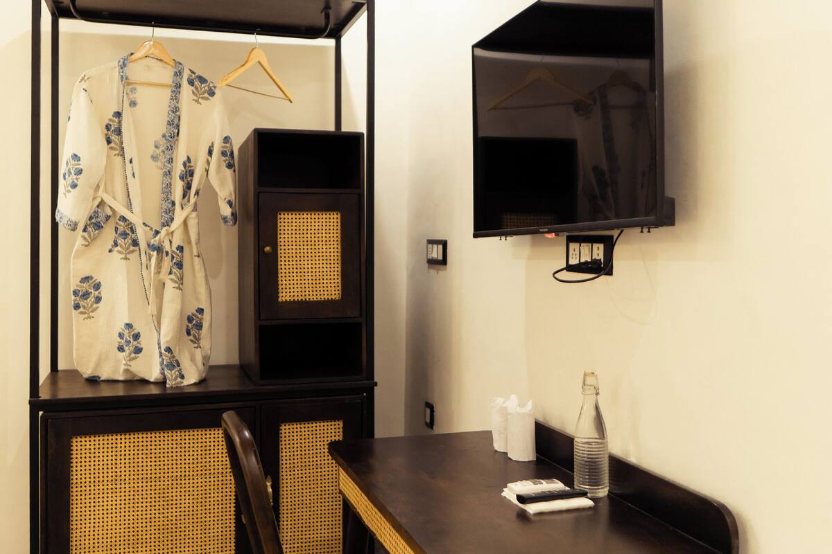 Una habitación compacta con cama, una mesita con una silla, televisor montado en la pared y una planta en maceta.
