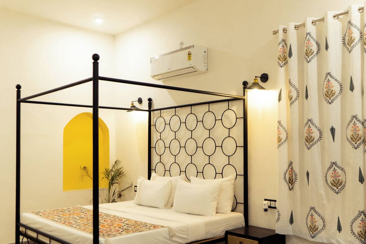 Una cama con dosel con cabecera adornada con diseños circulares, un ventilador de techo, un equipo de aire acondicionado, una mesita de noche y una silla.