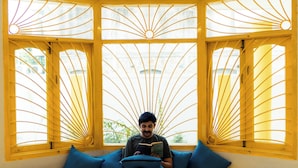 Eine Person sitzt auf einer gelben Bank mit blauen Kissen und liest ein Buch. Der Raum hat Fenster mit gelben Rahmen und einen Marmorboden.
