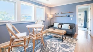 Ein Wohnzimmer mit Sofa, zwei Sessel, einem Couchtisch und einer Lampe. Es gibt große Fenster und einen Deckenventilator.