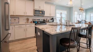 A modern kitchen with white cabinets, a large island with bar stools, a microwave, and a hanging light fixture. - Lone Star Breeze (Carolina Beach)