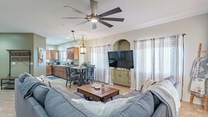 A living room with a sofa, coffee table, and TV, connected to a kitchen with wooden cabinets and a dining area. - Luxe Top-Floor Condo|Camelback Views| Heated Pool (Scottsdale)