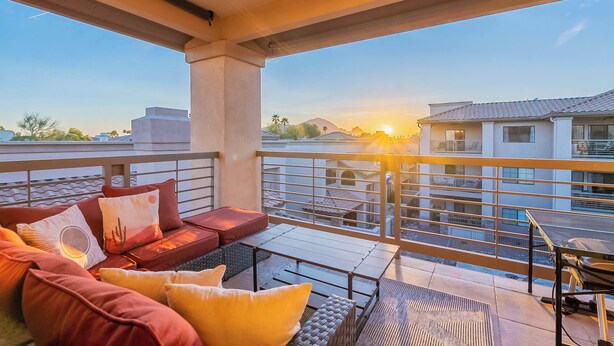 A balcony with a sofa, a table, and a view of buildings and a sunset. - Luxe Top-Floor Condo|Camelback Views| Heated Pool (Scottsdale)