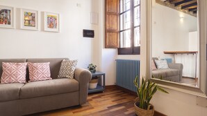 A living room with a sofa, a small table with a plant, a radiator, and a window with a wooden shutter. - Giardini 47 - Morada (Cagliari)