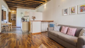 A cozy living area with a wooden floor, a sofa with patterned cushions, a kitchen with a wooden countertop, and a dining area with a table and chairs. - Giardini 47 - Morada (Cagliari)