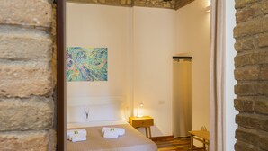 A hotel room with a bed, a nightstand with a lamp, a bathroom with a shower curtain, and a painting on the wall. - Giardini 47 - Morada (Cagliari)