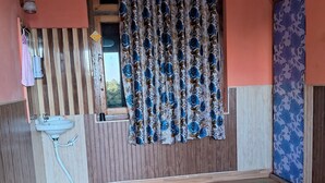 Une pièce avec un plancher en bois, un lavabo, une fenêtre avec des rideaux et une porte recouverte d’un papier peint à motifs floraux.