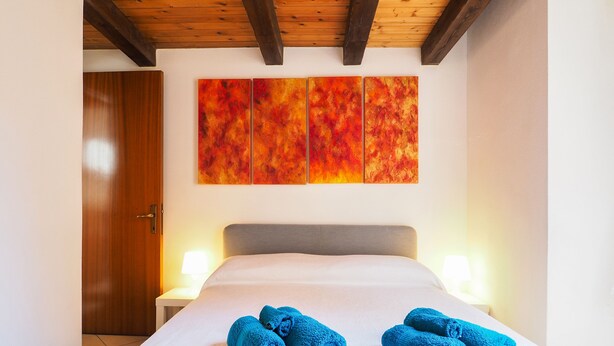 Une chambre d’hôtel avec un plafond en bois, un lit recouvert de draps blancs, deux lampes de chevet et un grand tableau abstrait accroché au mur.