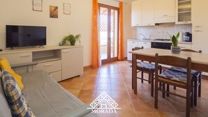 Un espacio habitable compacto con sofá, una mesa de comedor con sillas, zona de cocina con armarios y estufa, y un televisor sobre un mueble.