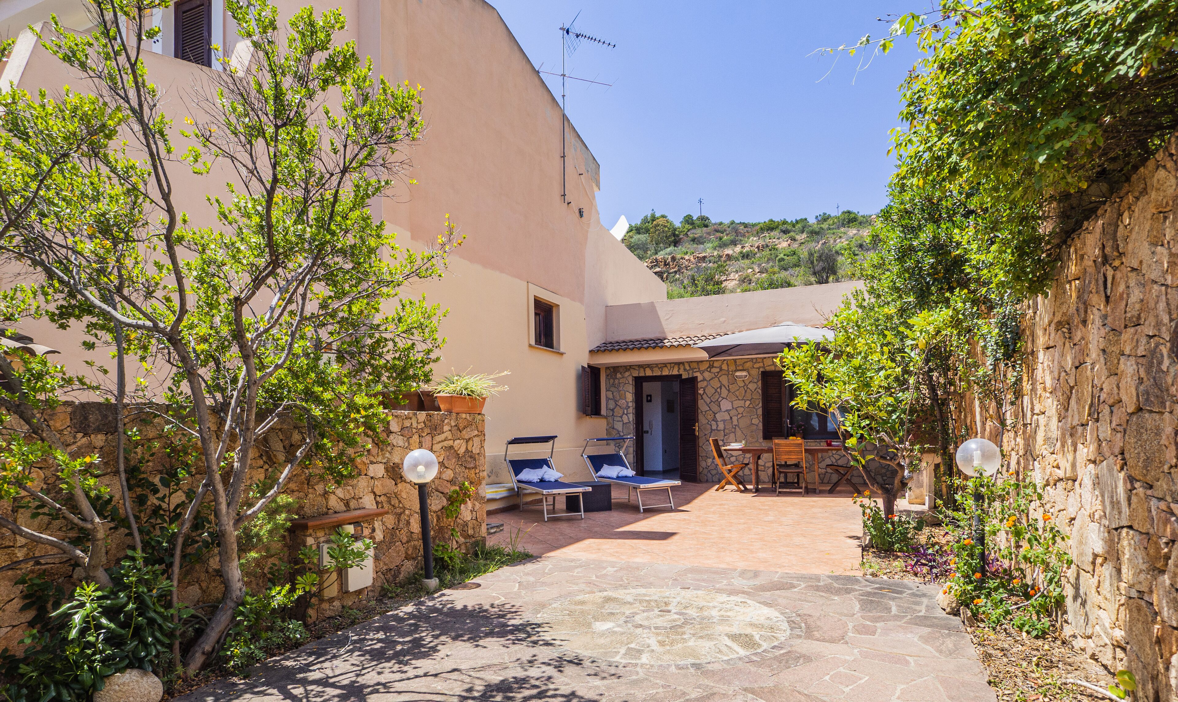 A stone pathway leading to a patio with two lounge chairs and a table, surrounded by greenery and a stone wall.