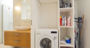 Ein kleines Badezimmer mit einem Holzkosmetikschrank, einem Waschbecken, einer Whirlpool Waschmaschine und einem Regal mit Reinigungsmitteln.