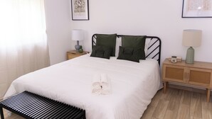 Un lit bien fait, avec une couette blanche et des serviettes pliées, deux tables de chevet avec des lampes et des œuvres d’art encadrées accrochées aux murs.