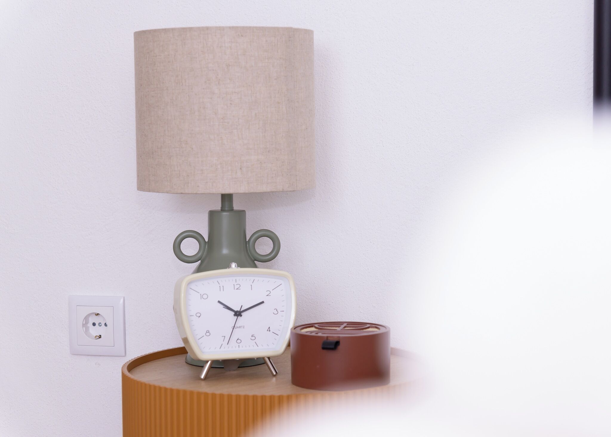 Una lámpara de pared con pantalla color beige, un reloj de estilo antiguo y un pequeño recipiente marrón sobre una superficie de madera.