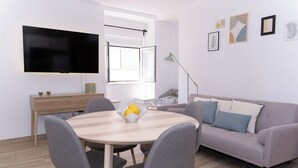 Un salon moderne avec une table à manger ronde, des chaises grises, un canapé gris, un téléviseur à écran plat, une fenêtre avec des stores ouverts et quelques œuvres d’art accrochées au mur.