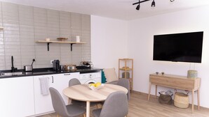 Une cuisine moderne avec des armoires blanches, un comptoir noir et une table à manger en bois avec quatre chaises. Un téléviseur est fixé au mur et une petite étagère en bois se trouve au-dessus du comptoir.