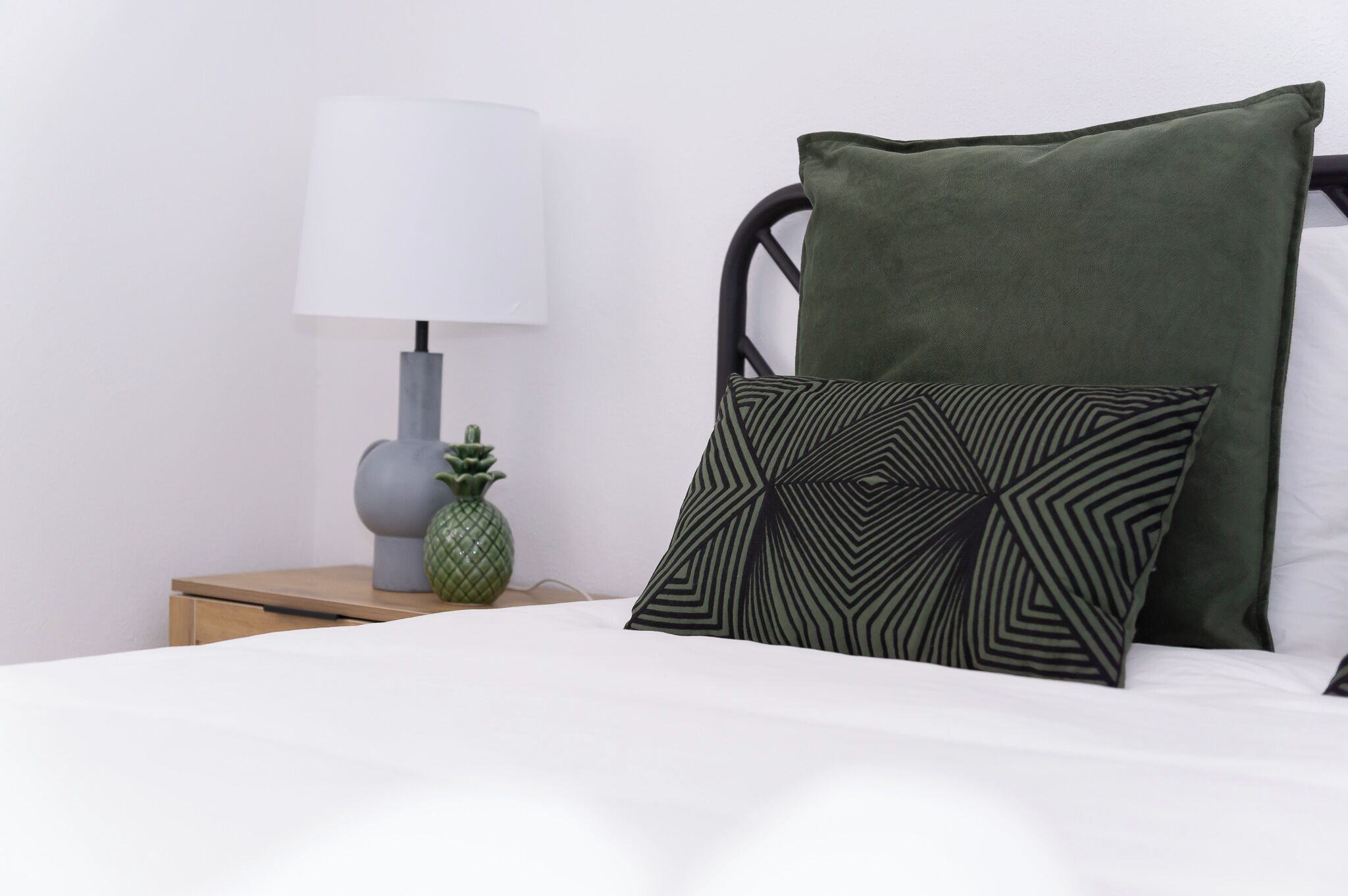 Una cama bien tendida con un almohadón verde y otro con estampado geométrico, una lámpara de noche y una pequeña decoración de piña sobre una mesita de madera.