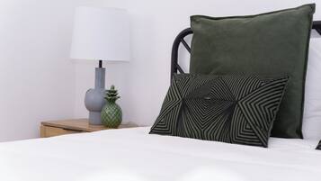 Una cama bien tendida con un almohadón verde y otro con estampado geométrico, una lámpara de noche y una pequeña decoración de piña sobre una mesita de madera.