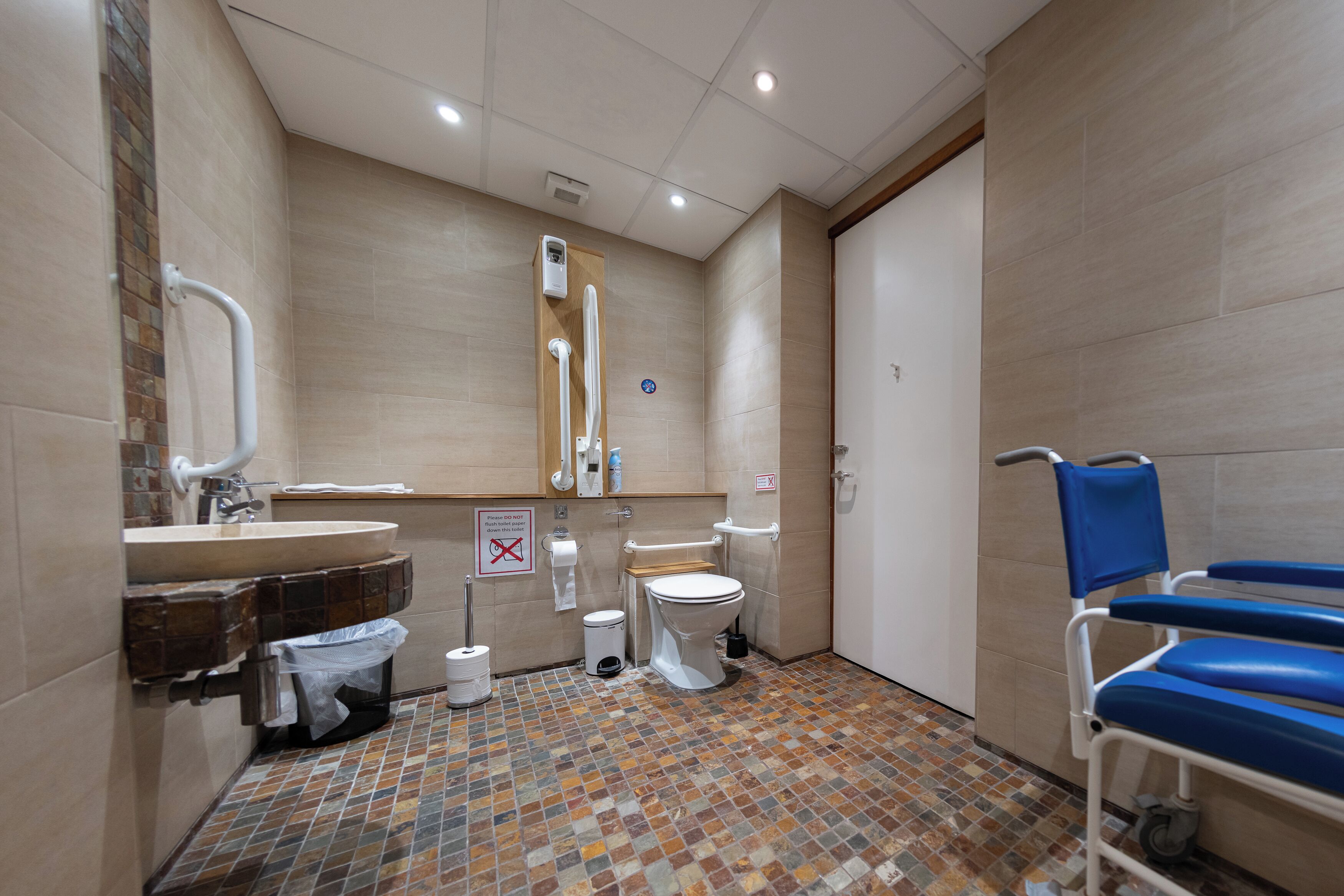 A bathroom with a tiled floor, a sink, a toilet, a bidet, and a wheelchair-accessible chair.