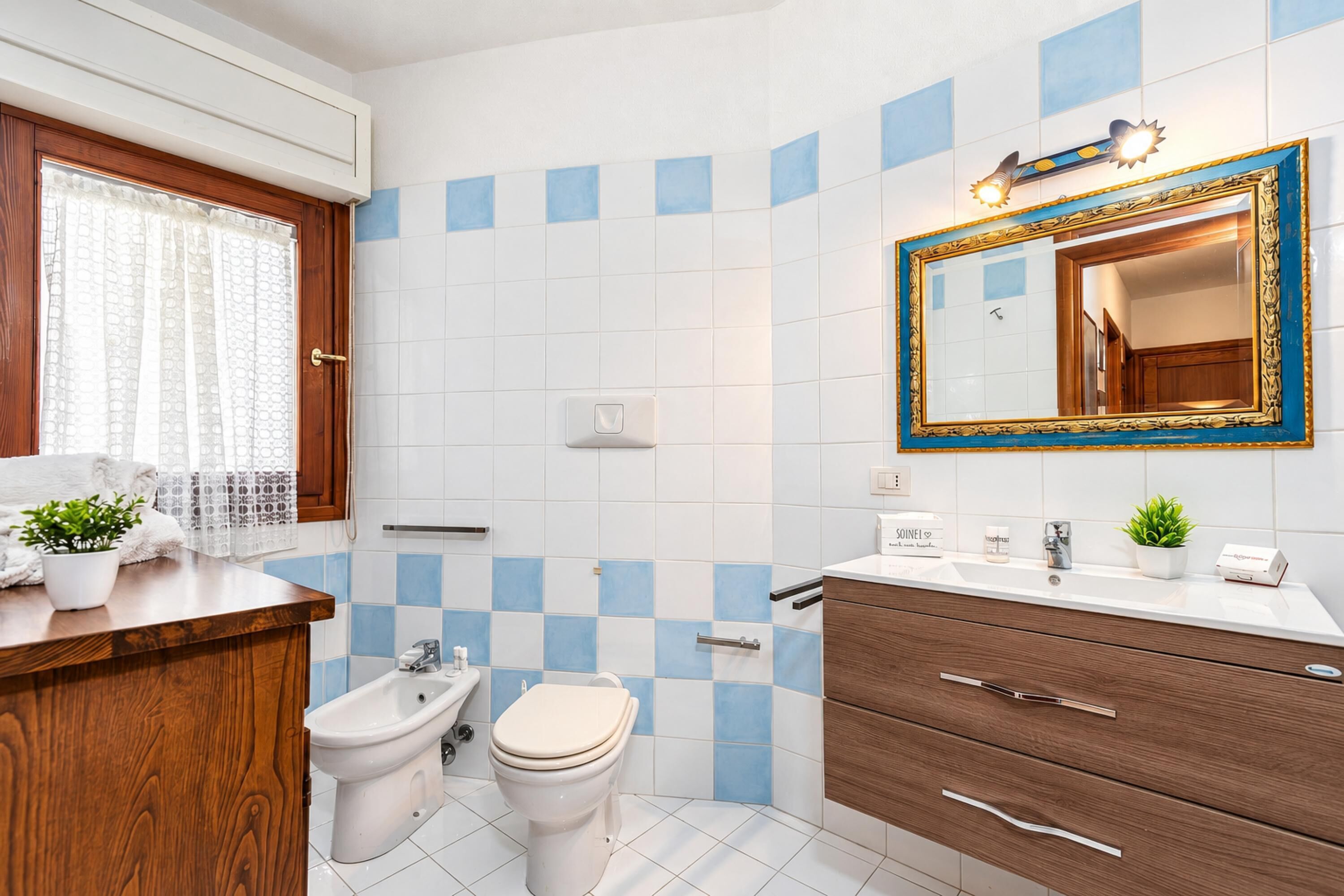 A bathroom with a bidet, toilet, vanity with a mirror, and a window with curtains.