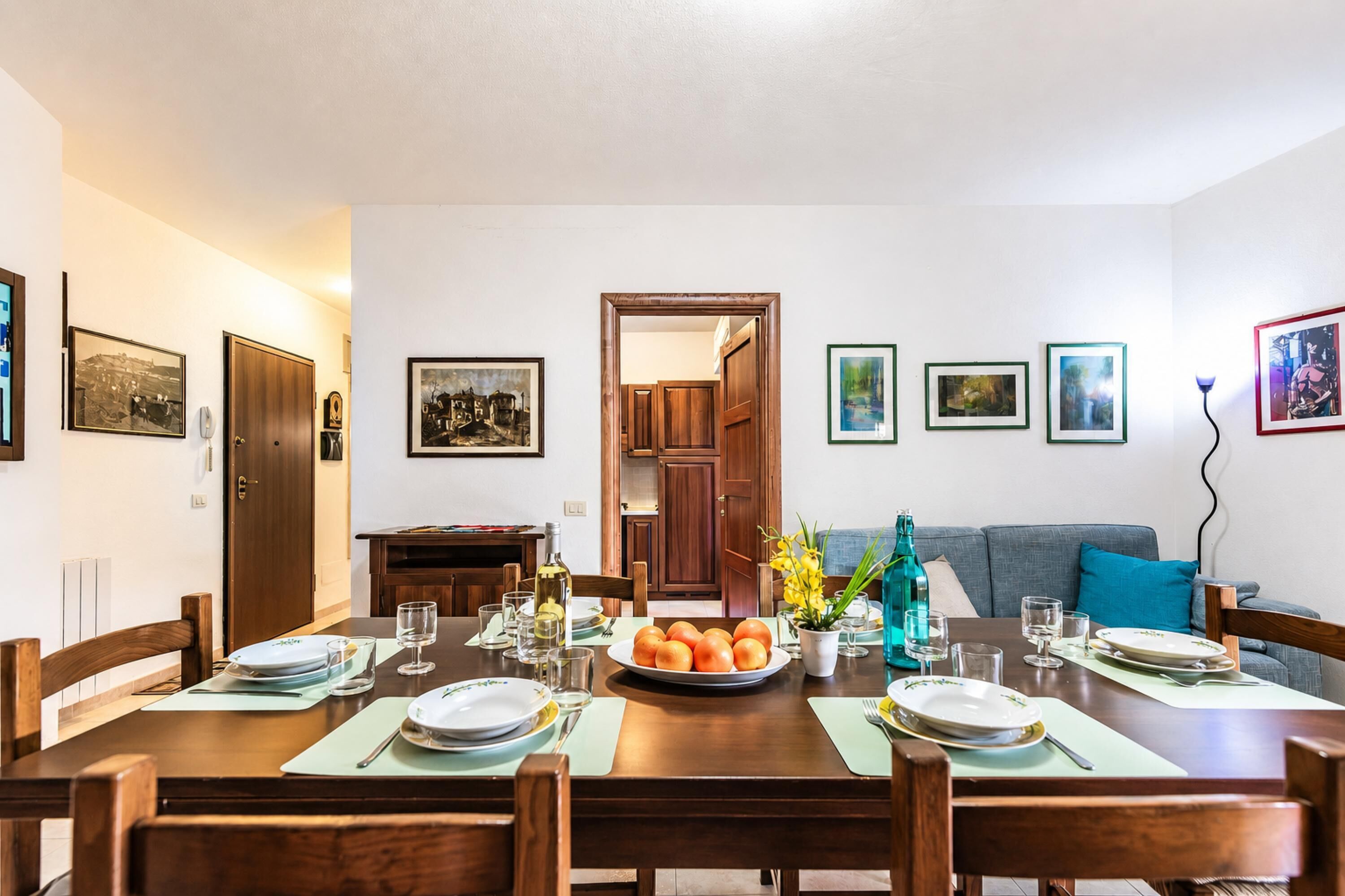 Uma sala de jantar com uma mesa de madeira posta para quatro pessoas, rodeada por cadeiras. Há quadros nas paredes, um sofá ao fundo e uma porta que dá para outro quarto.
