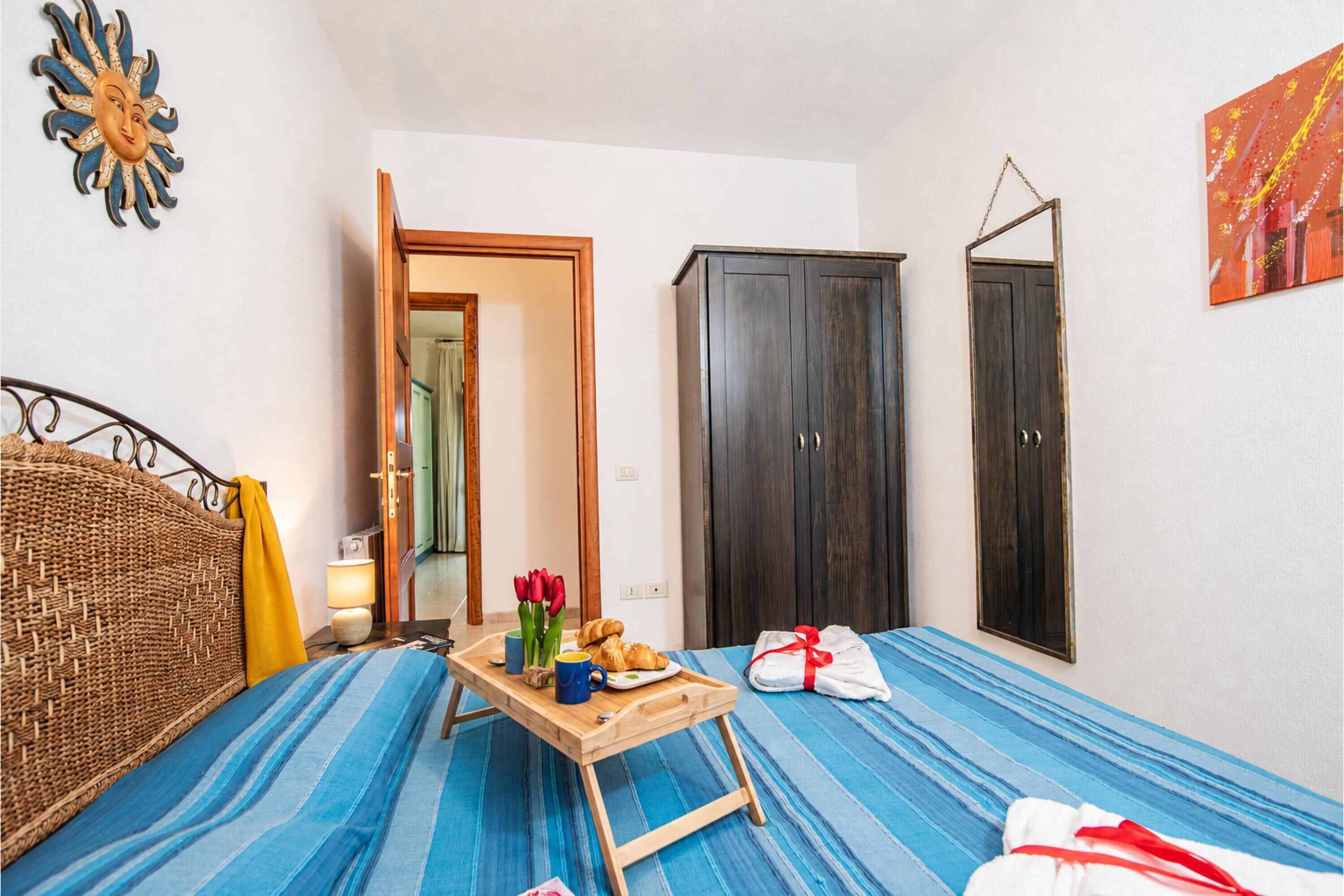 A room with a bed, a wooden table with items, a wardrobe, and a mirror.
