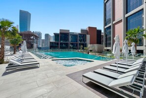 Uma área de piscina no terraço com espreguiçadeiras e guarda-sóis, rodeada por edifícios modernos e palmeiras.