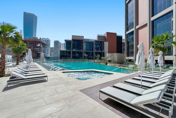 A rooftop pool area with lounge chairs and umbrellas, surrounded by modern buildings and palm trees.