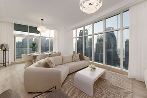 A modern living room with a large sectional sofa, a coffee table, and a dining area with a table set for four. Large windows offer a city view.