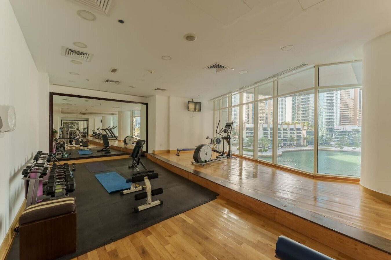 Une salle de sport spacieuse, équipée de matériel d’exercice, dotée d’un grand miroir et offrant une vue sur le paysage urbain grâce à de larges fenêtres.