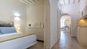 Een slaapkamer met een bed, nachtkastje en dressoir. Er is een gang die leidt naar een keuken met kasten en een aanrecht.