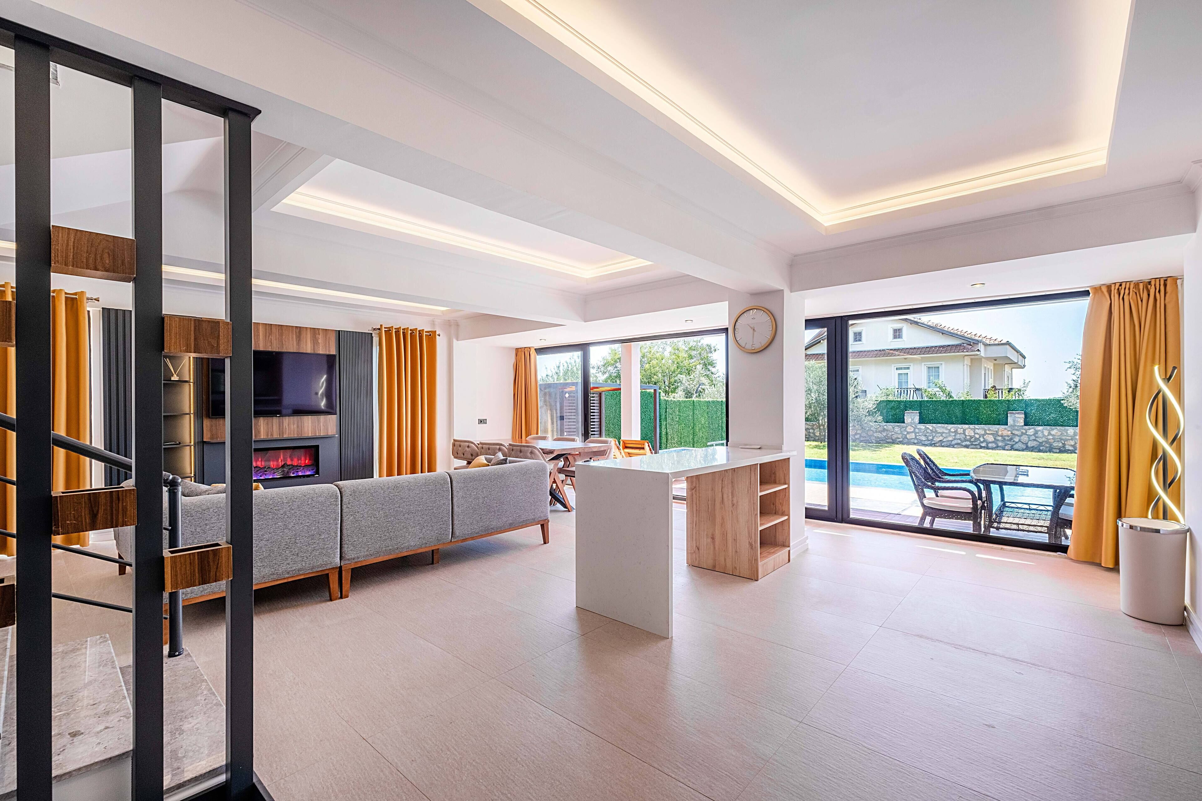 Amplia sala de estar con sofá, televisión y chimenea. Hay una mesa de comedor con sillas y vistas a una piscina exterior.