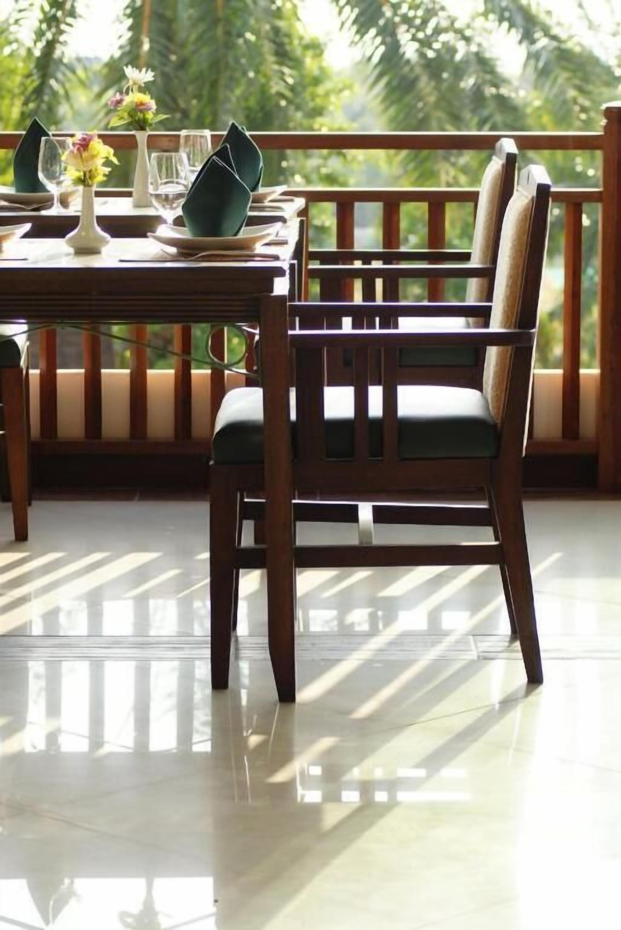 A dining area with a wooden table and chairs, a vase with flowers, and a view of greenery through a balcony railing.
