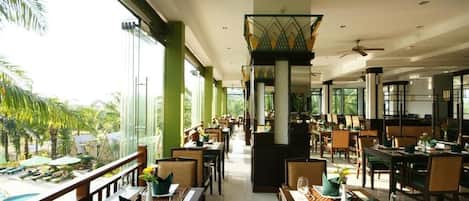 A spacious restaurant with high ceilings, large windows, and a view of palm trees. Tables are set with green napkins and cutlery.