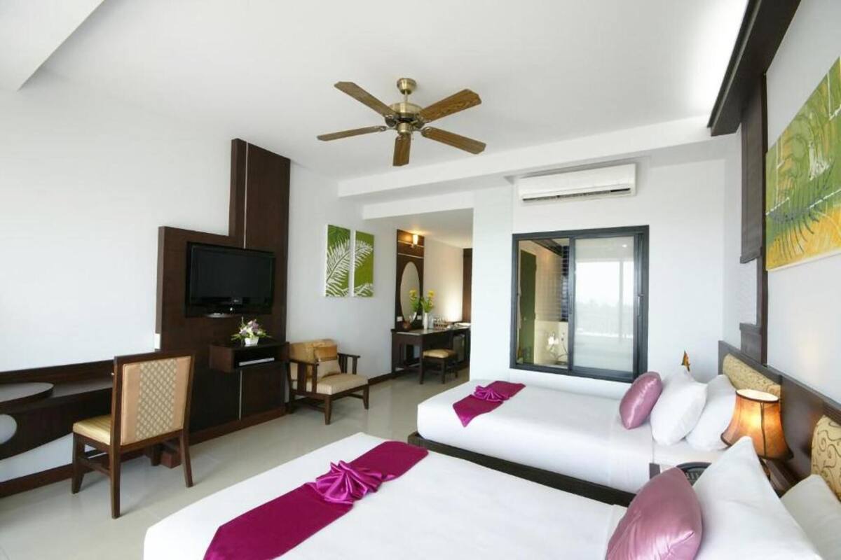 A hotel room with two beds, a desk, a television, and a ceiling fan.