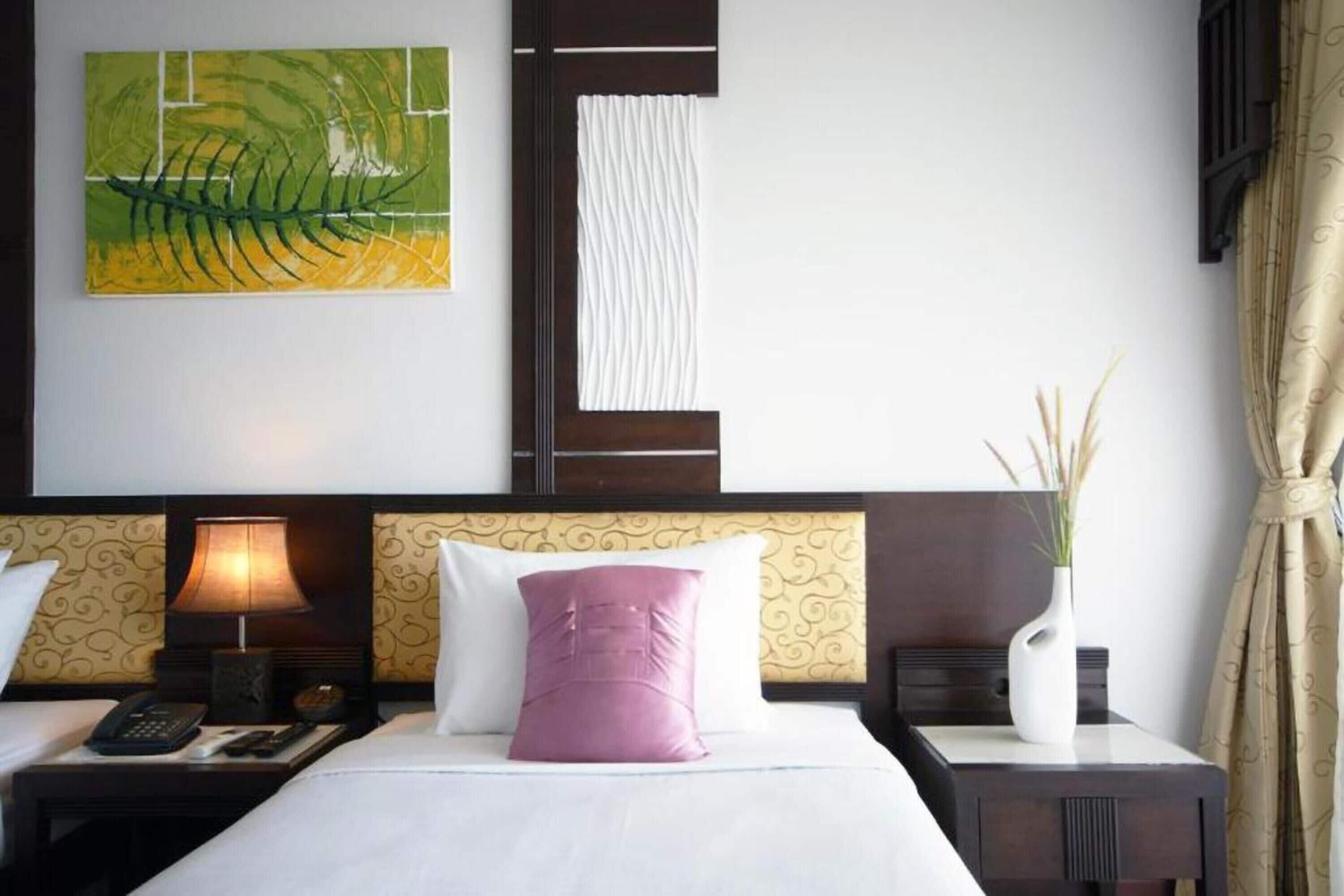 A hotel room with a bed, two bedside tables, a lamp, a telephone, a decorative painting, a vase with plants, and a curtain.