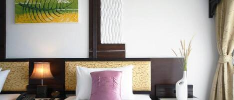 A hotel room with a bed, two bedside tables, a lamp, a telephone, a decorative painting, a vase with plants, and a curtain.