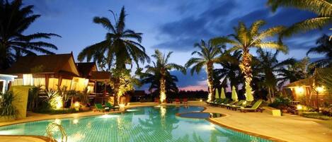 A swimming pool with palm trees and lounge chairs, surrounded by buildings with warm lighting.