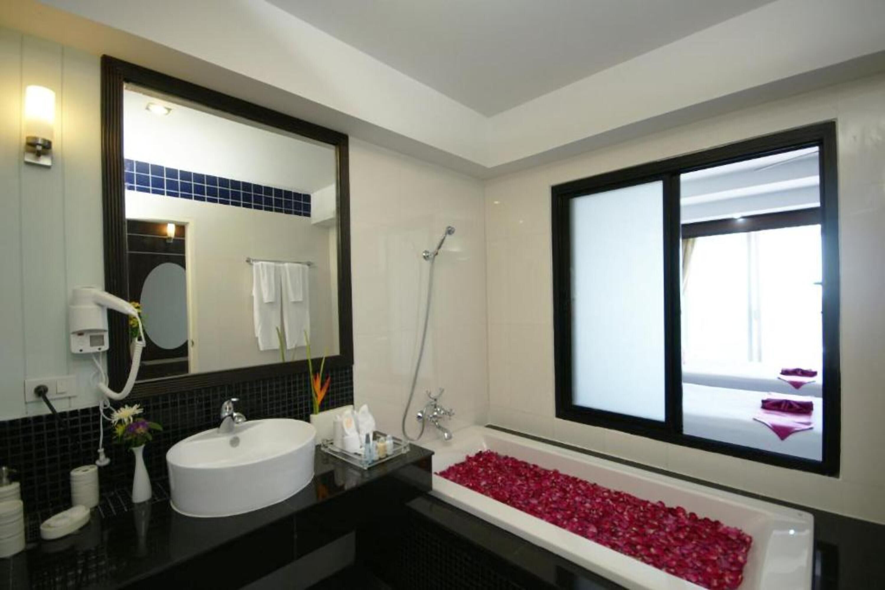 A modern bathroom with a white sink, a large mirror, a bathtub filled with rose petals, and a window providing natural light.