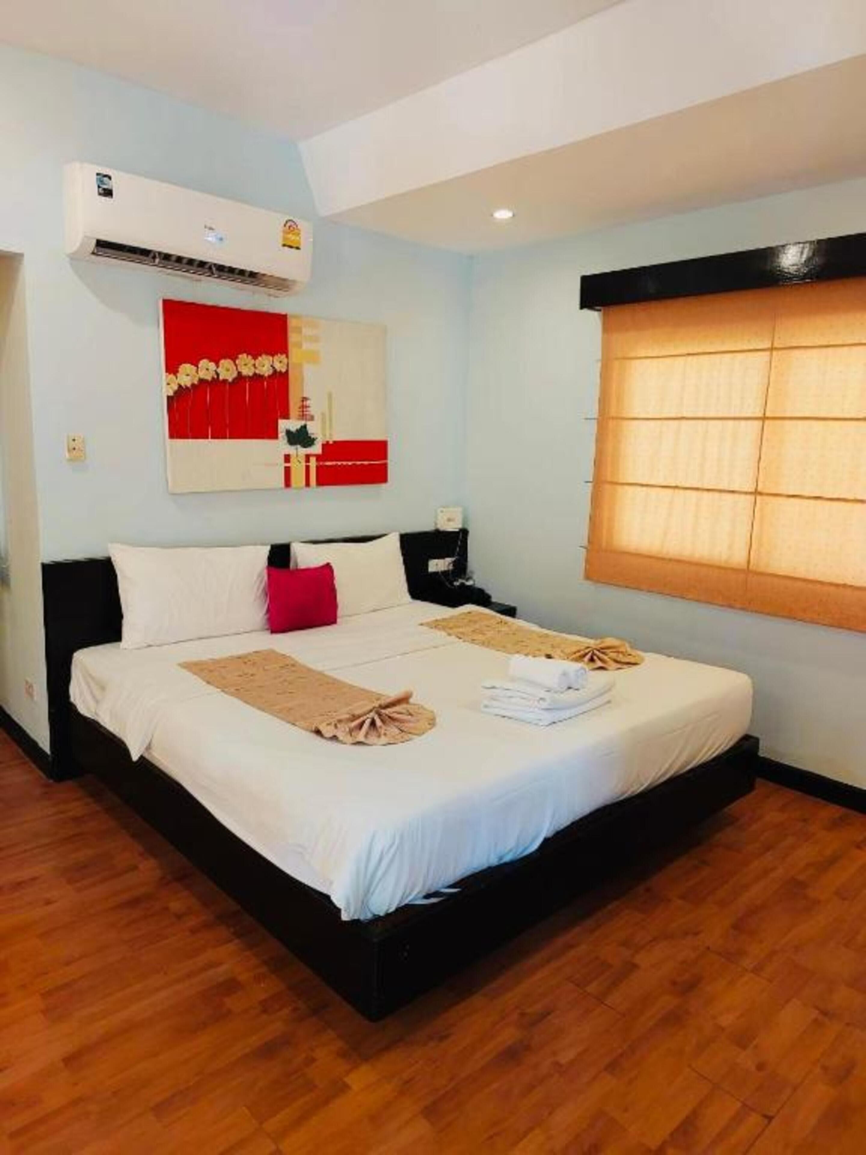 A neatly made bed with a red pillow, a white pillow, and a beige throw blanket. There is a wall-mounted air conditioner and a painting above the bed.